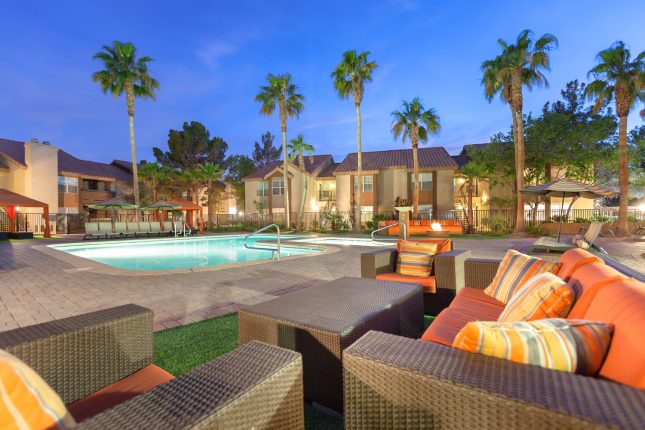 An apartment complex swimming pool at dusk with wicker furniture with orange cushions in the foreground and palm trees and apartments in the background.