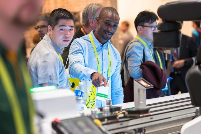An African-American in a blue shirt man pointing to an item on an assembly line beside an Asian man at the Bosch trade show exhibit at the Consumer Electronics Show.