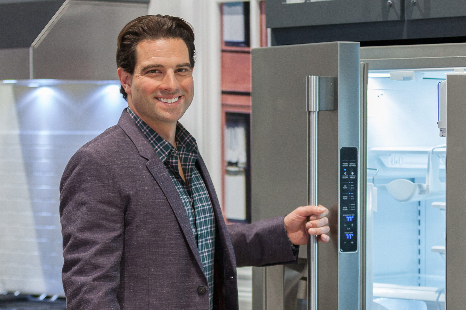 HGTV's Scott McGillivray, a white male smiling in a grey suit jacket opening a stainless steel refrigerator with illuminated white interior in the KBIS 2016 Electrolux exhibit.
