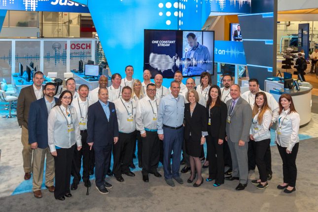 A team photograph of 25 on grey carpet in front of a blue and grey modern trade show exhibit.