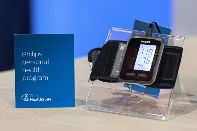 A close up of a Philips blood pressure monitor on a clear vinyl stand and a blue sign beside it upon a whitewashed wood surface.