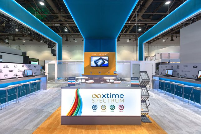 Trade show exhibit with three soaring blue ceiling elements above, an illuminated white welcome kiosk in the foreground and long counters with modern barstools on the right and left.