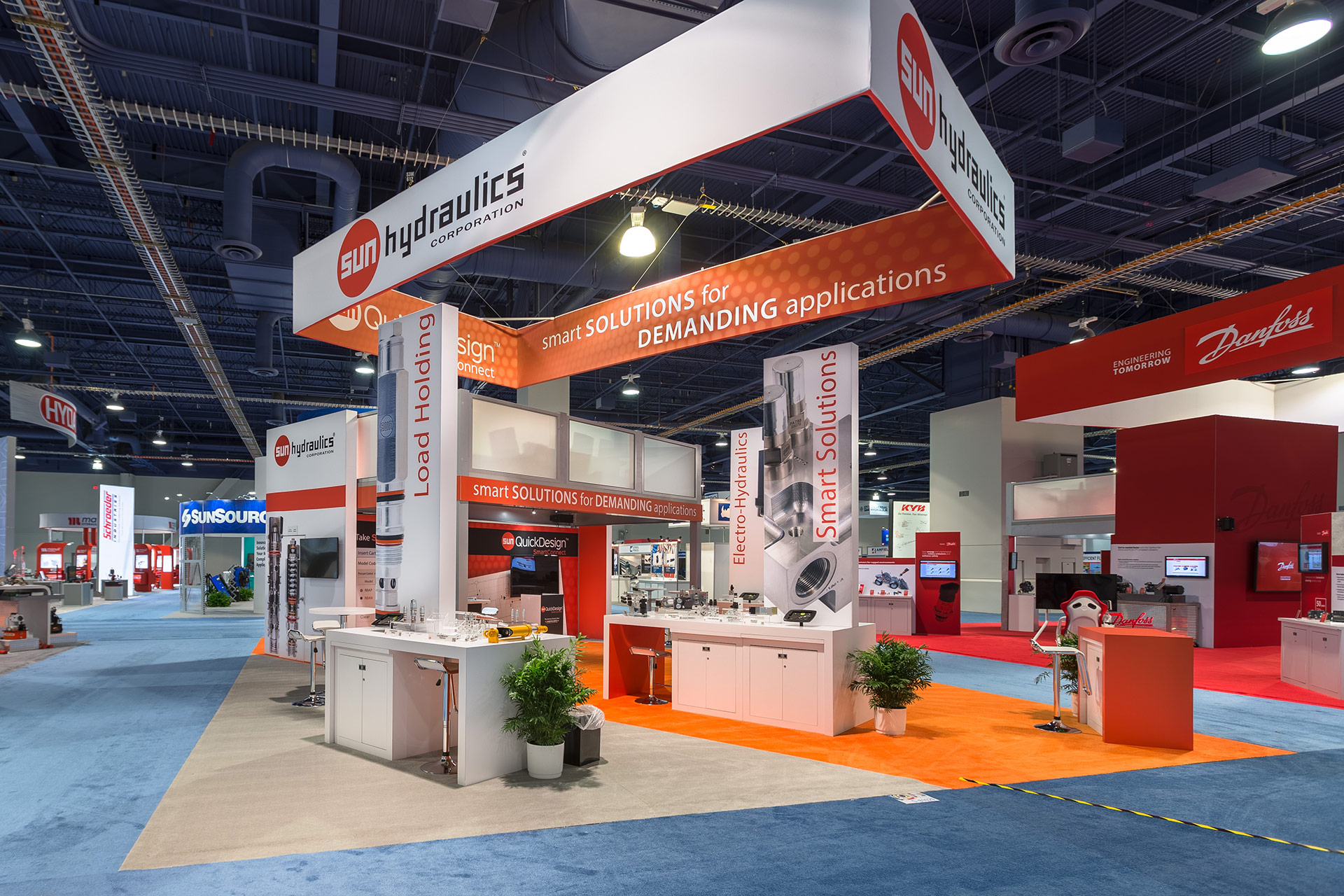 Trade show exhibit with an orange and white color scheme, a suspended rectangular banner with four faces above and multiple product displays within and a two-story platform in the background. Convention center has blue carpet and other exhibits are visible.