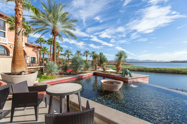 A hotel fountain pond surrounded by palm trees and and desert lake in the distance and a small table and two chairs in the foreground.