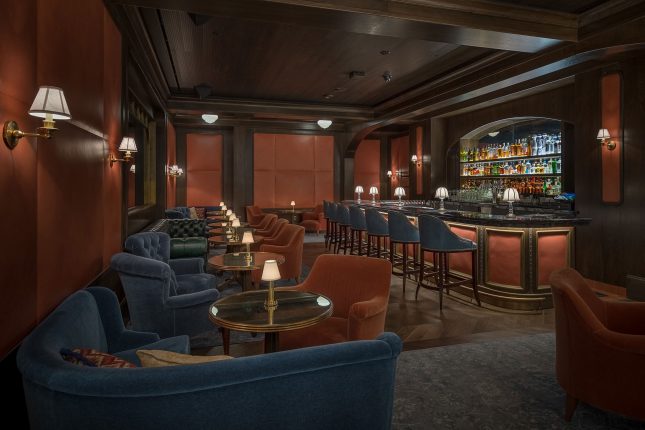 A dark lounge with cushioned orange walls and wall lamps, a wooden ceiling with a series of short drink tables bearing miniature lamps on them and orange and teal velvet chairs surrounding each as well as a bar with teal velvet barstools on the right.