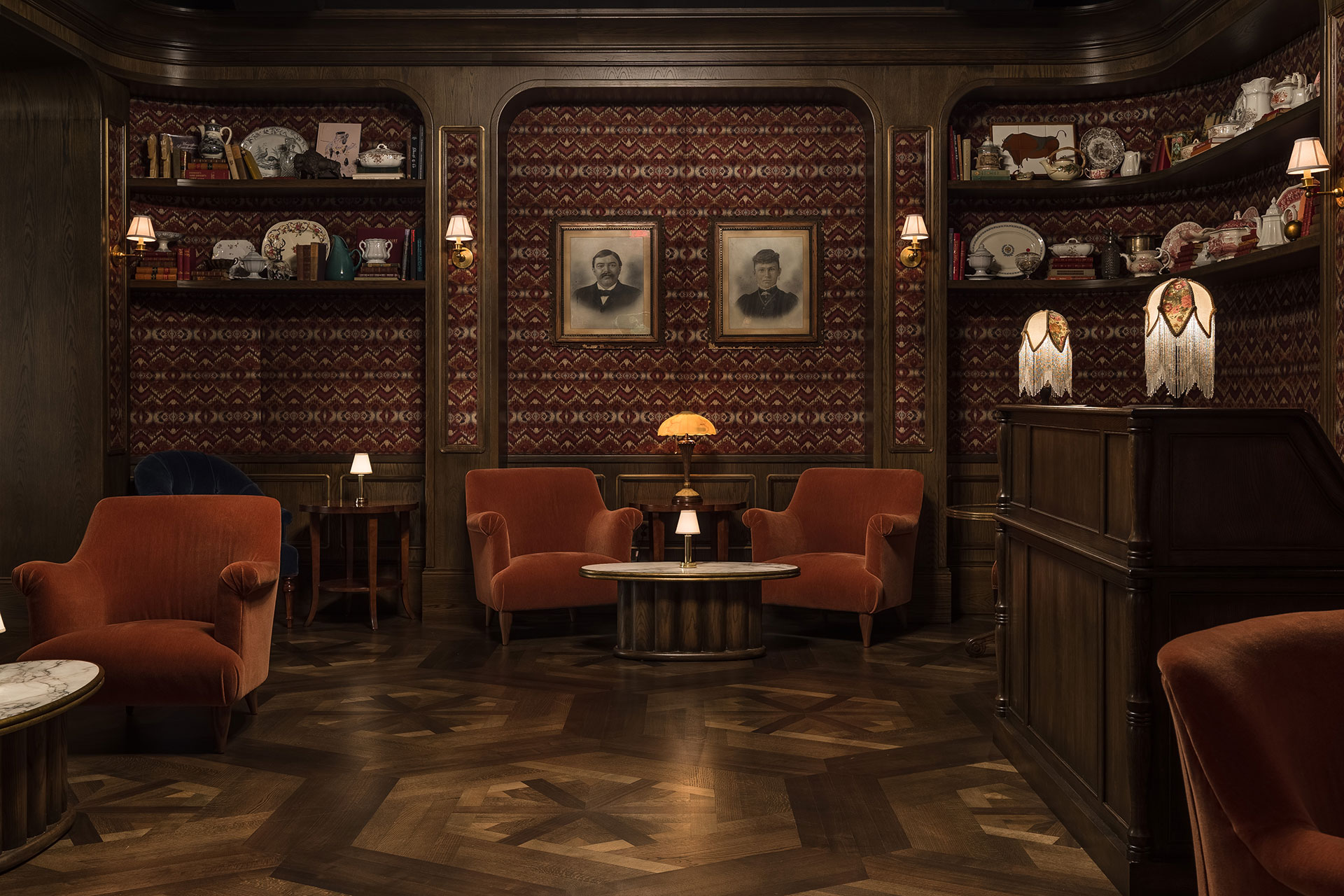 a dark, moody restaurant entry with custom woody flooring in a hex pattern, two velvet chairs, lamp, and drink table in the center with antiques on shelves above on a wallpapered wall and a tall wooden host's podium on the right.