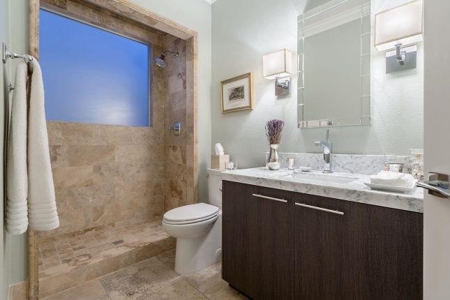 A bathroom with dark wood cabinet, white marble countertop and beige tile in bath with frosted window glowing blue with dusk light.