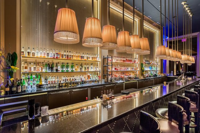 A row of lampshades with tassles above a long black bar with purple-seated barstools and a wall of liquor bottles in the background.