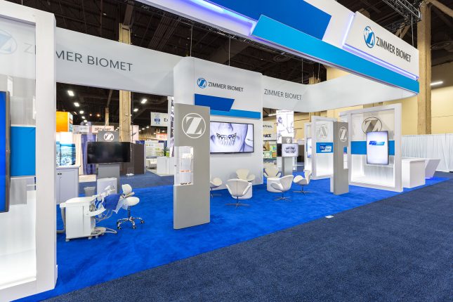 A Trade Show Exhibit with blue carpet, tall and narrow, grey and white vertical display cases, white chairs, and suspended signage.