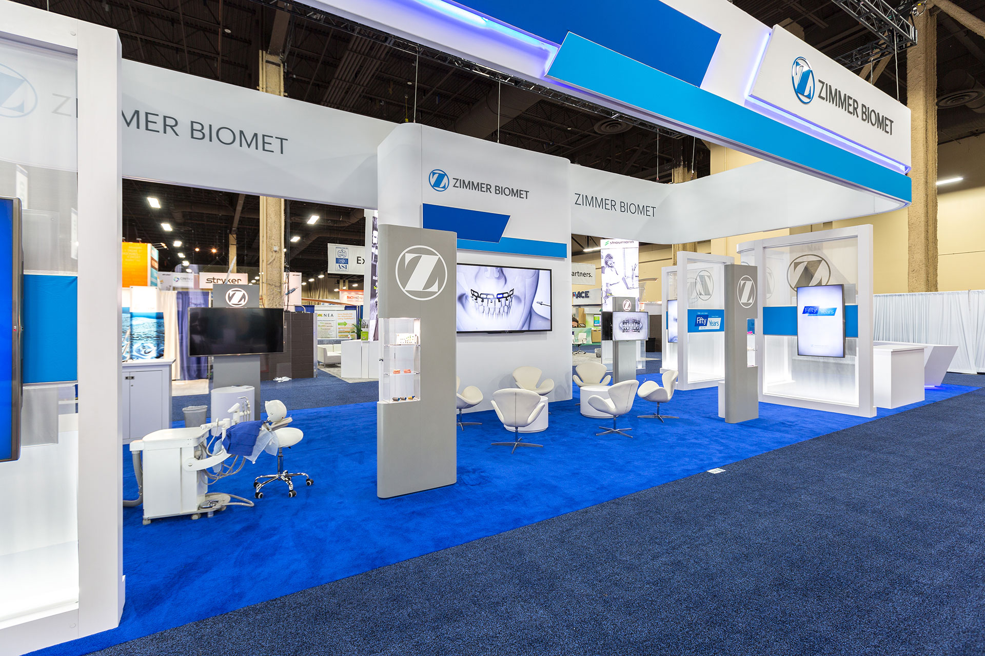 A Trade Show Exhibit with blue carpet, tall and narrow, grey and white vertical display cases, white chairs, and suspended signage.