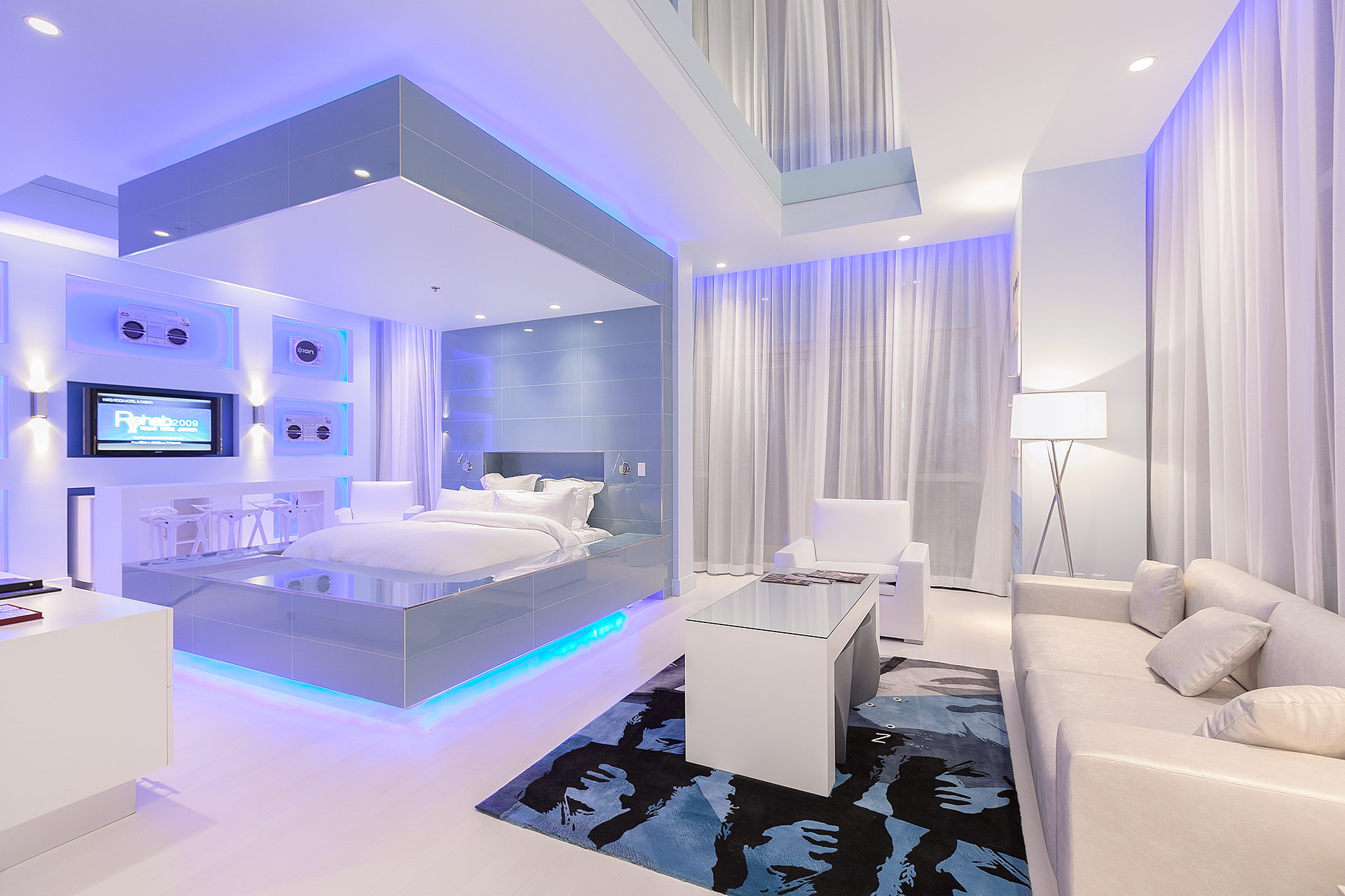 A modern white hotel room with a c-frame bed with blue led lighting above, below, and illuminating sheer, white drapes with a blue-and-black graphic-art rug in the foreground and blue, led-lit niches in the background with jam-boxes mounted in them.