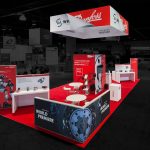 An elevated perspective of a Danfoss White Drive trade show exhibit with red carpet, white tables and counters with multiple red vertical displays throughout and a suspended banner with their logo above and a pony wall with a black and blue print of a gear in the foreground. Photo edited so that the outside area is darkened and colorless.