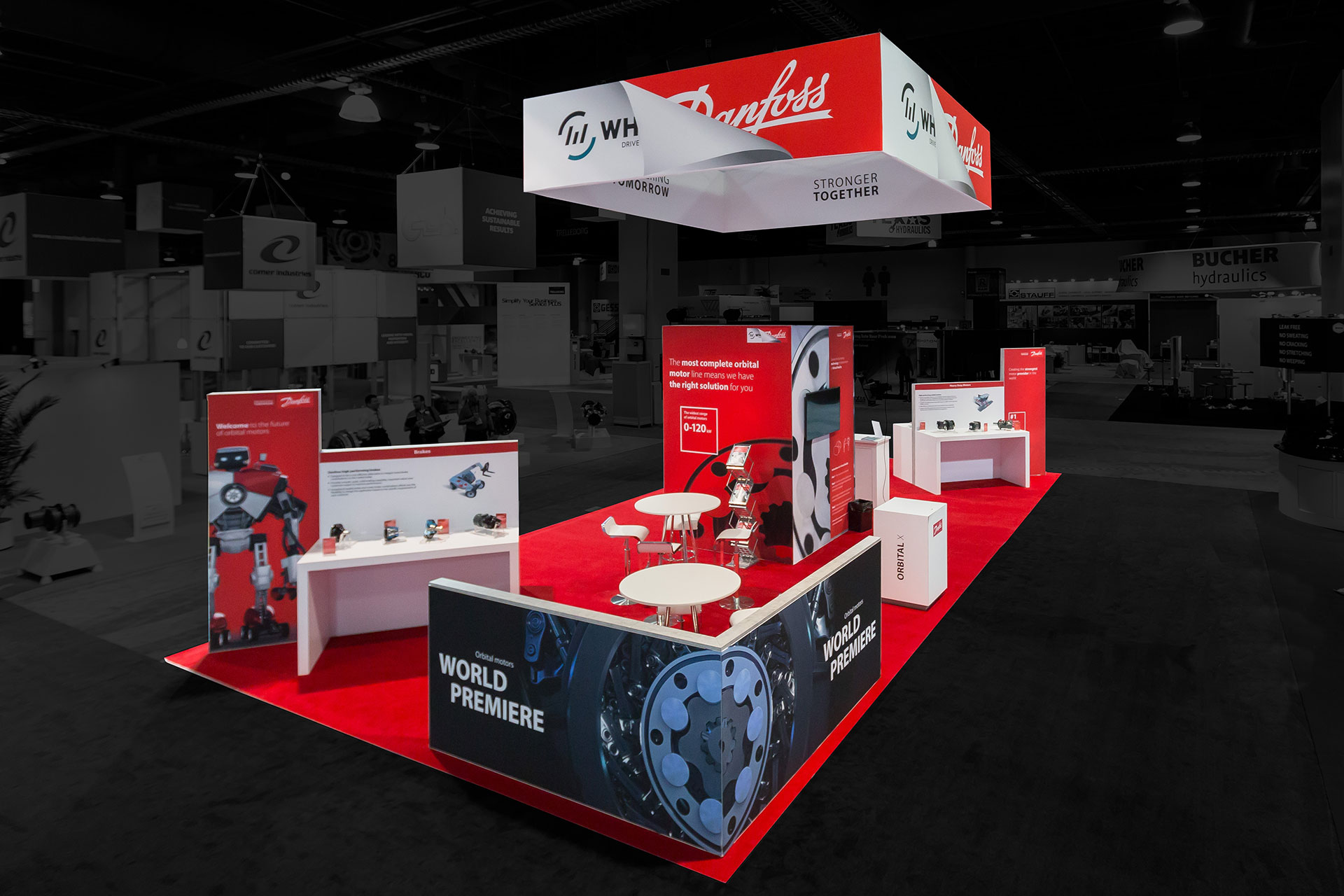 An elevated perspective of a Danfoss White Drive trade show exhibit with red carpet, white tables and counters with multiple red vertical displays throughout and a suspended banner with their logo above and a pony wall with a black and blue print of a gear in the foreground. Photo edited so that the outside area is darkened and colorless.