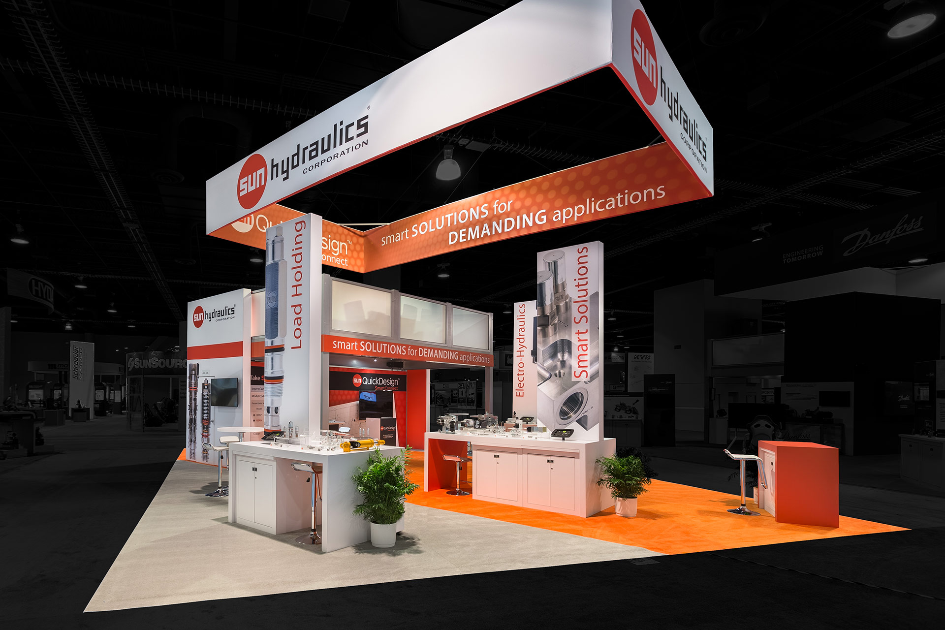 Trade show exhibit with an orange and white color scheme, a suspended rectangular banner with four faces above and multiple product displays within and a two-story platform in the background. Photo edited so that the outside area is darkened and colorless.