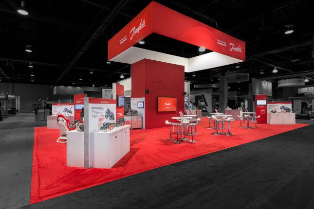 A trade show exhibit from an oblique angle with a red wall in the center, rectangular red banner floating above and red carpet with modern white tables, chairs and kiosks edited so that area outside the booth is darkened and colorless.