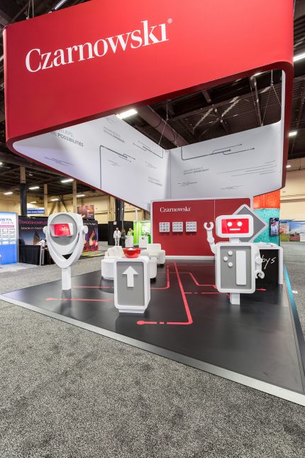 A red, white, and black booth with robot-looking video displays and a large suspended sign with inlaid text.