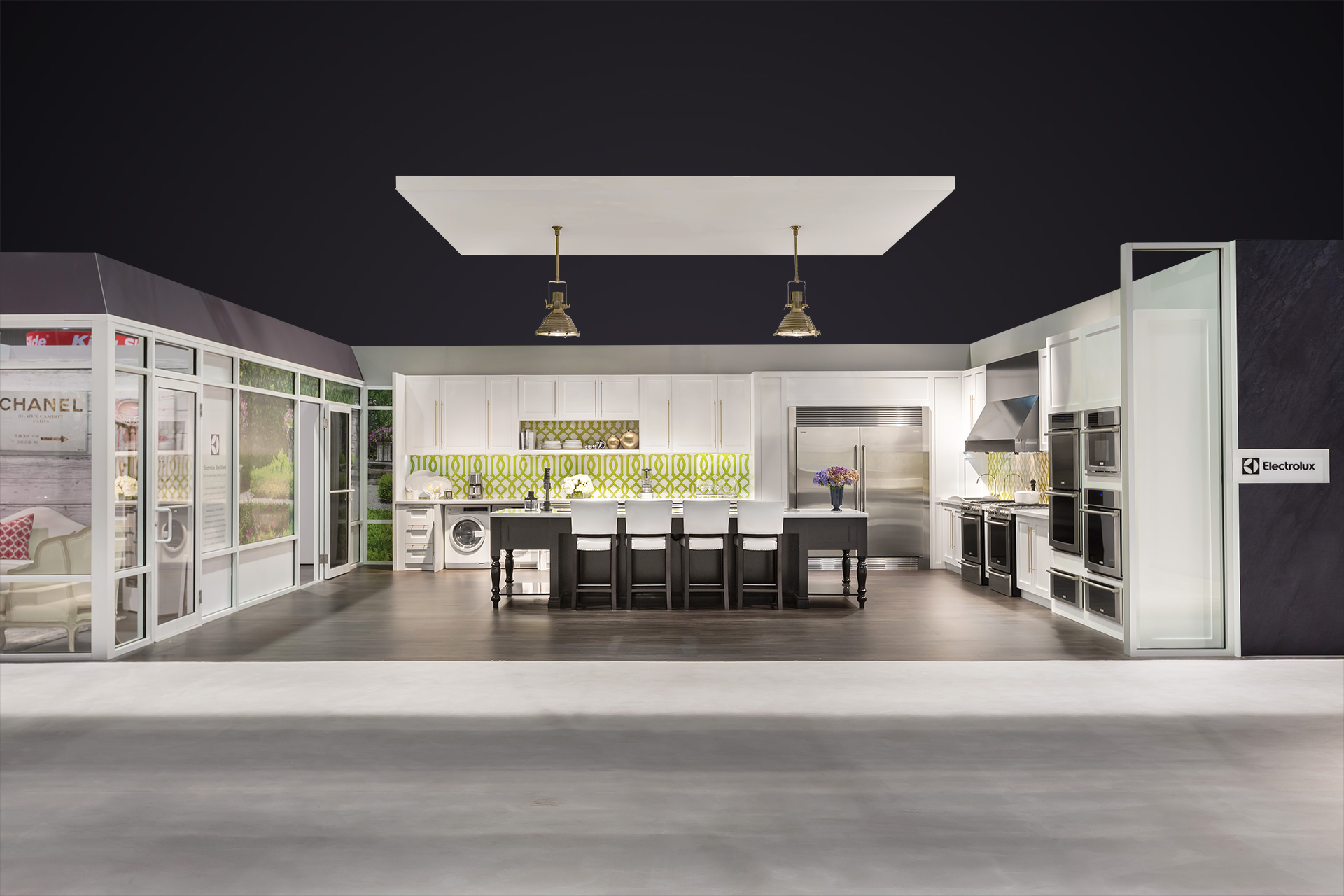 A wide angle of the Electrolux tradeshow exhibit kitchen with white cabinets stainless steel appliances and a black wooden island with a suspended soffit and two gold hanging lamps over greyish brown hardwood floors and below a purple digitally edited ceiling.
