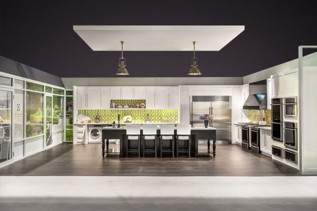 A tradeshow exhibit kitchen with white cabinets stainless steel appliances and a black wooden island with a suspended soffit and two gold hanging lamps over greyish brown hardwood floors and below a purple digitally edited ceiling.