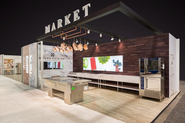 A kitchen trade show exhibit with a stainless steel cooking island in the foreground and a cooking station behind it with a ten-foot wide video screen, dark brown hardwood wall on one side and marble wall with two video screens on the other side beneath copper pots and pans and the word "Market" suspended above.