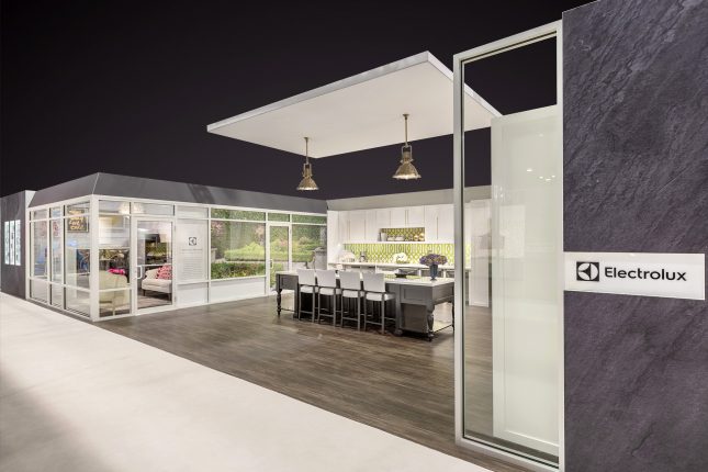 A grey wall in the foreground with the Electrolux logo and a kitchen island on a dark hardwood floor beneath a suspended ceiling and two lamps with white cabinetry in the background and a glass wall with two doors on the left.