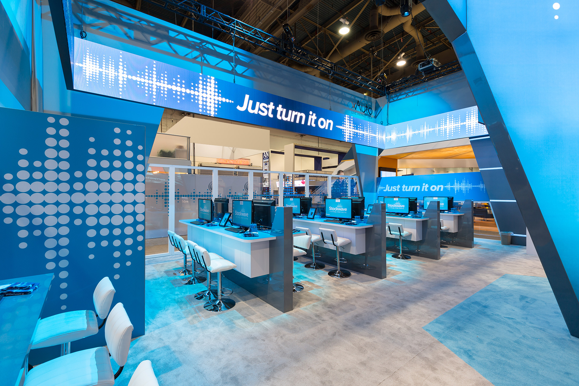 Four rows of white counters under blue lighting with white padded barstools and video monitors displaying a blue Stockwave logo beneath a thin video wall which surrounds the exhibit displaying the text "just turn it on".