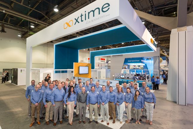 A group of 30 people in blue shirts and khaki pants in front of a trade show exhibit with a suspended white ceiling with the xTime logo above.