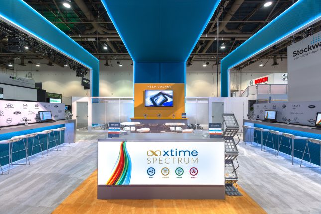 Trade show exhibit with three soaring blue ceiling elements above, an illuminated white welcome kiosk in the foreground and long counters with modern barstools on the right and left.