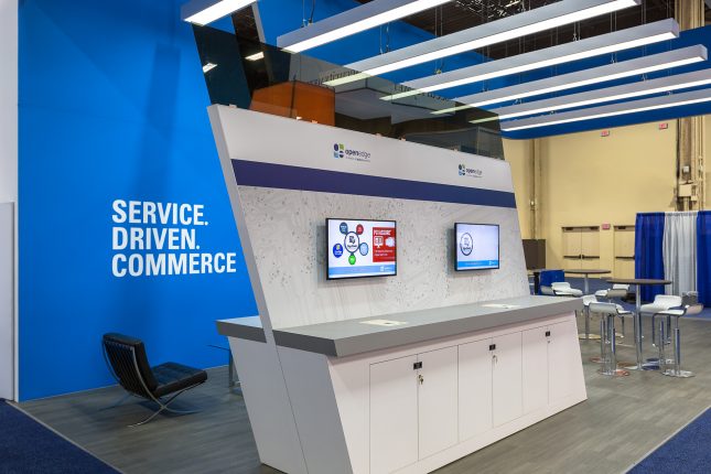 A tall parallelogram-shaped kiosk with two video monitors and grey counter space in the foreground with a blue wall in the background, a black barcelona chair on the left and tall tables and barstools on the right and long, thin fluorescent light fixtures above.