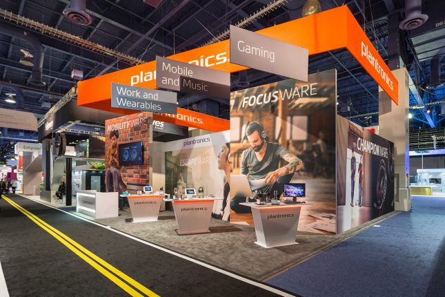 Three white Plantronics display tables with a video monitor and products atop in the foreground, with printed walls of young people using laptops and headphones in the background and hangind printed signage above.