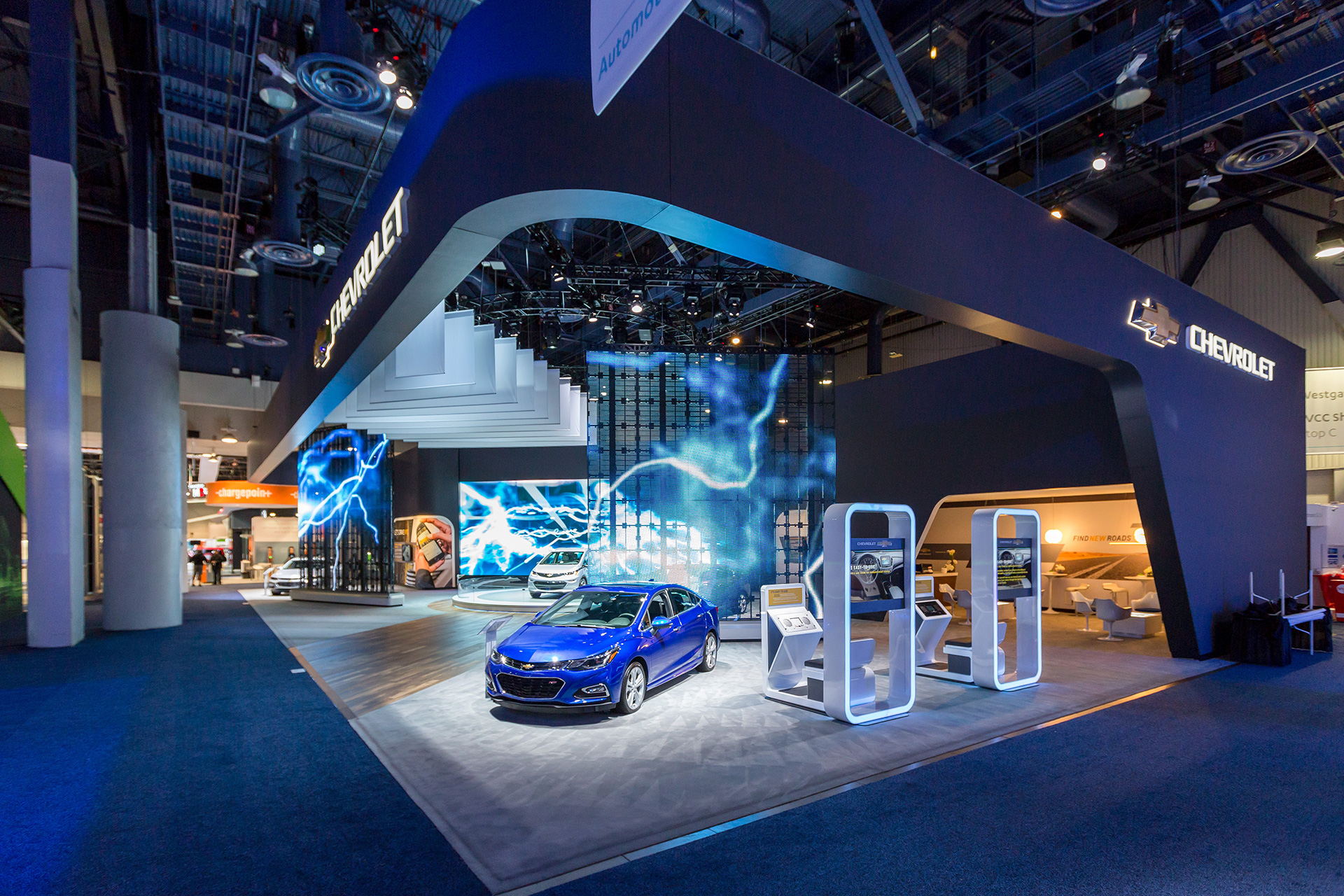 Chevrolet exhibit with a blue Chevy volt in the foreground, rounded rectangular interactive kiosks, video walls with lightning, and a grey volt on a pedestal in the background.