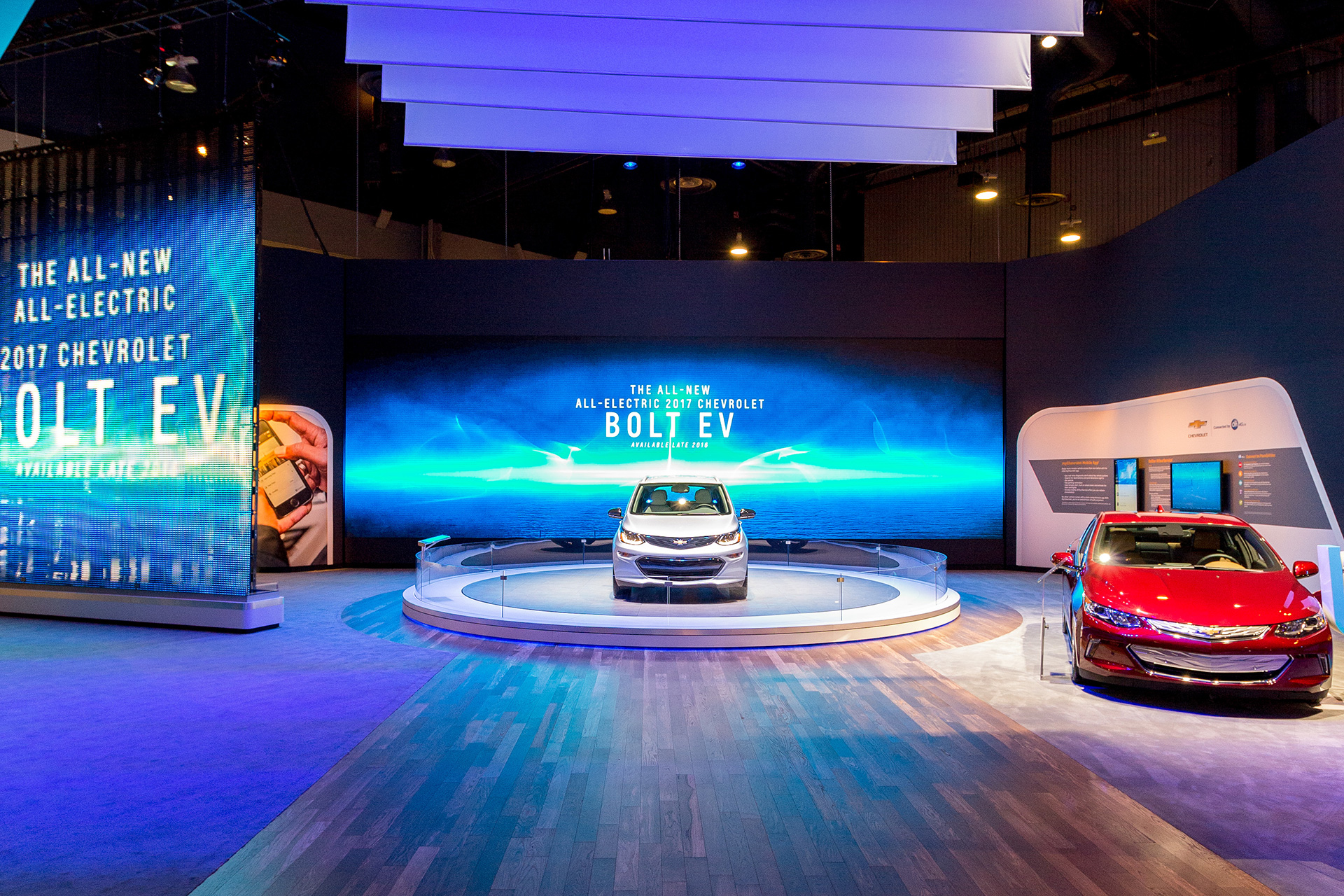 Hardwoode floor leading to a circular pedestal with a grey chevy bolt atop, a a red vehicle to the right and a video wall with ethereal imagery and text in the background.