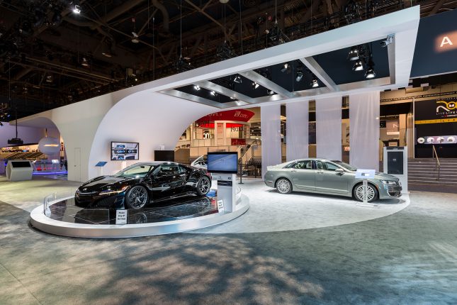 A grey car on the right and a black sports car on the left upon a circular pedestal on light an dark grey carpet with a arched white suspended ceiling and lighting inside a convention center.