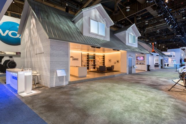 Exhibit shaped like a home with faux-white brick and gabled windows as an exterior multiple interior areas with a living room with two chairs and shelves on a wall in the foreground.