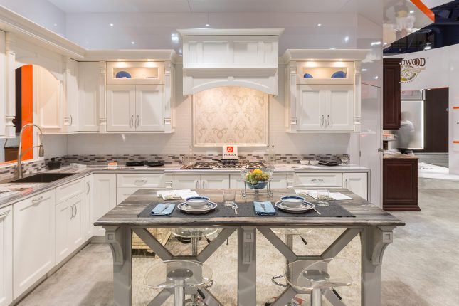 Tradeshow exhibit of white kitchen cabinets a grey island with marble-looking surface and tablesetting atop.
