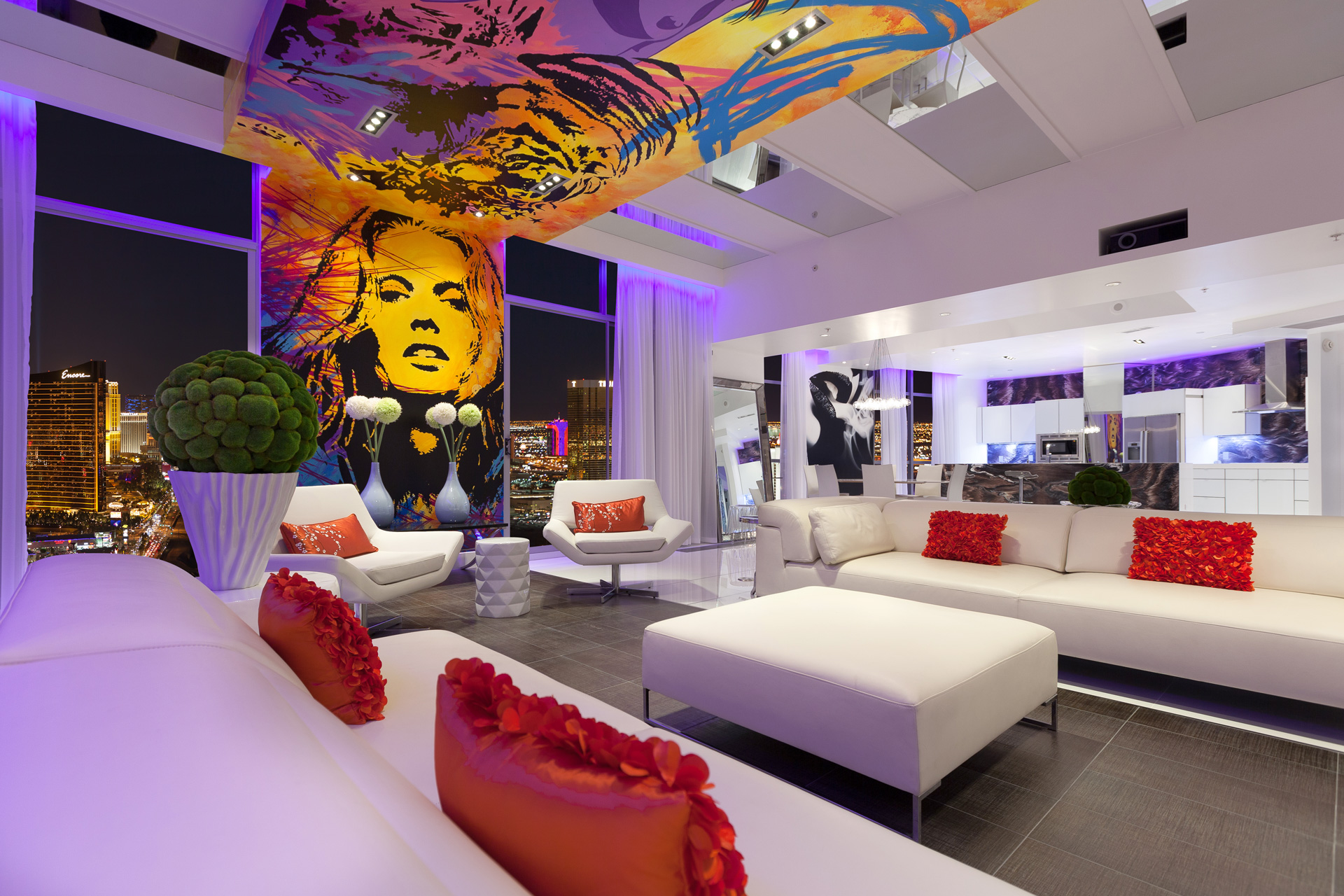 A chic las vegas penthouse with modern white seating, city views, colorful wall murals of women and a modern kitchen in the background.