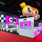 A black and white tradeshow exhibit with three floating inflatable powerpuff girls suspended above and a pink Cartoon Network logo on video screens.