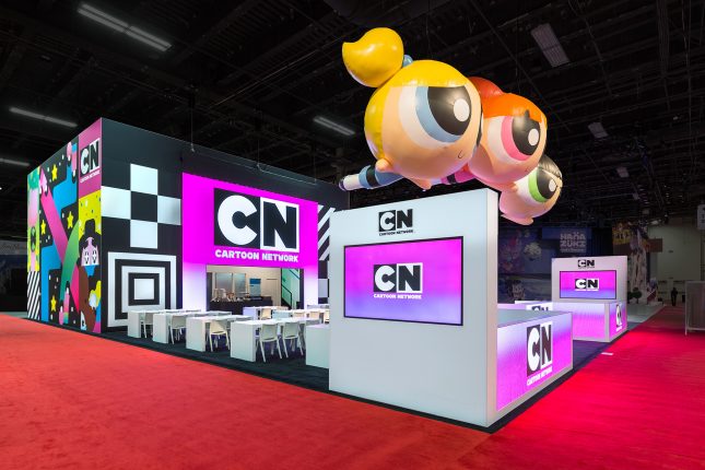 A black and white tradeshow exhibit with three floating inflatable powerpuff girls suspended above and a pink Cartoon Network logo on video screens.