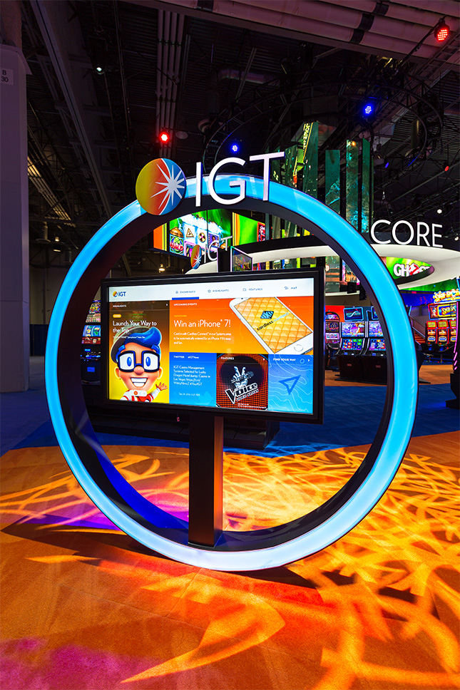 An illuminated circular frame with an interactive video screen inside and the IGT booth in the background at G2E.