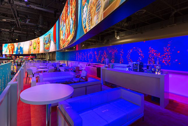 Sofas and tables on an elevated deck beneath a curved video wall at the IGT booth at G2E.