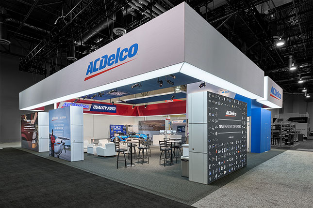 An angular view of the ACDelco tradeshow booth with a suspended grey banner and graphic blocks as walls with barstools, tables, and displays inside.