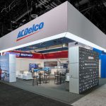 An angular view of the ACDelco tradeshow booth with a suspended grey banner and graphic blocks as walls with barstools, tables, and displays inside.