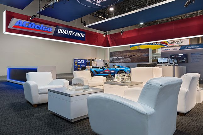 White sofa seating facing tables with auto parts encased in transparent cubes in the foreground with a red suspeded banner above emblazoned with the ACDelco logo and the words "Quality Auto".
