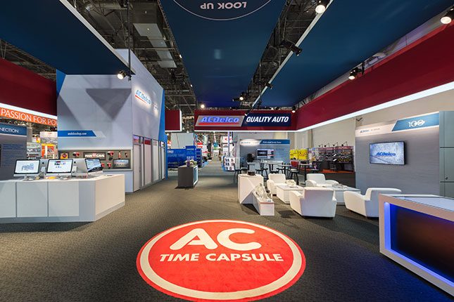 The inside of the ACDelco tradeshow booth with a red circular medallion on the floor, white seating to the right, computer kiosks on the left and in the rear the ACDelco logo suspended on a banner.