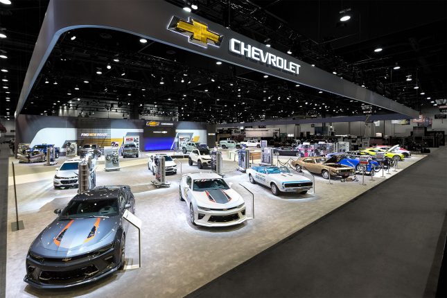 An elevated angular view of the Chevy booth at SEMA with classic Chevy Camaros lined up in the foreground and the Chevrolet logo emblazoned on a dark grey banner floating above.