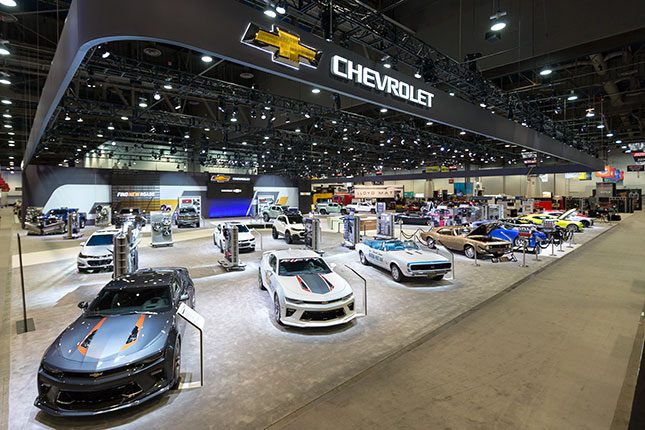 An elevated angular view of the Chevy booth at SEMA with classic Chevy Camaros lined up in the foreground and the Chevrolet logo emblazoned on a dark grey banner floating above.