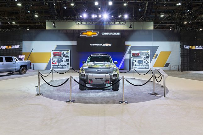 A prototype chevy utility vehicle behind stantions in a grey circular carpeted area with a video screen, two vertical displays with Chevy parts attached and the chevrolet logo in the background.