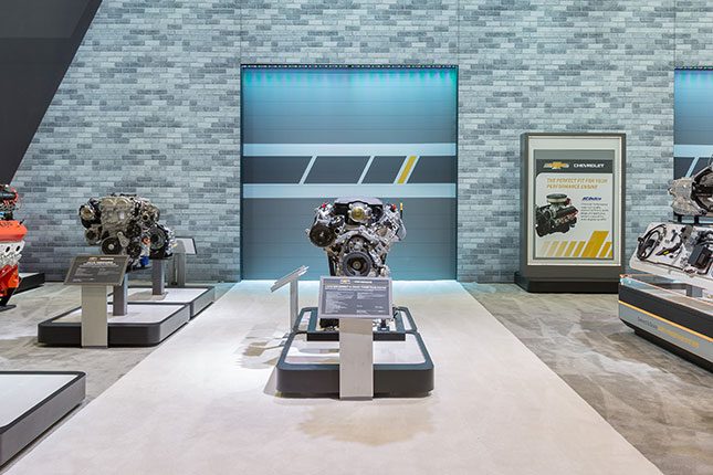 A Chevy engine on a pedestal atop tradeshow carpeting with a faux brick wall and garage door in the background.