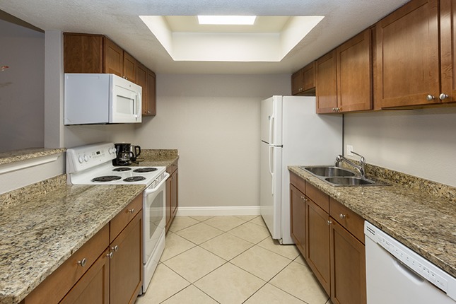 An apartment kitchen with tan tile brown grantie countertops a white range and microwave to the left and a stainless steel sink and white refrigerator to the left.