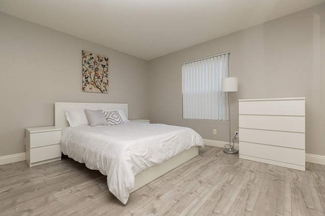 A bedroom with grey walls, hardwood floors, a bed to the left with white sheets and a lamp and white dresser to the left.
