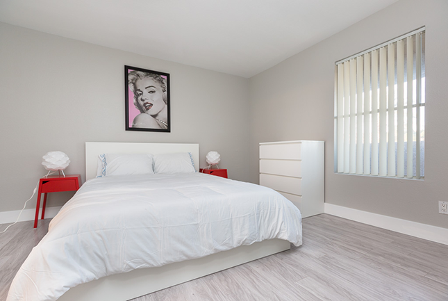 A bedroom with grey walls, grey hardwood, pop art of Marilyn Monroe hanging above a bed with white sheets and red nightstands with a white dresser and a vertical blinds of a window to the left.